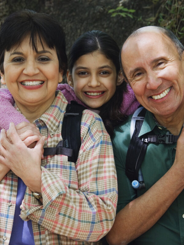 A family of three smiles into the camera. The mom is on the left and the father is on the right. The daughter poses between them.