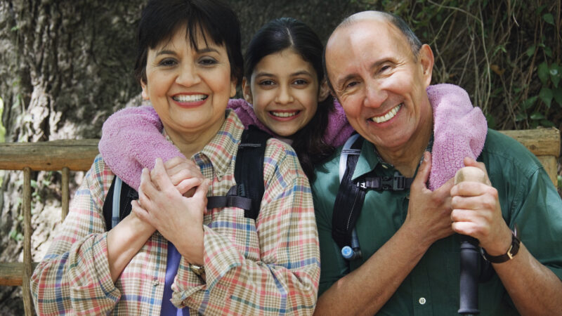 A family of three smiles into the camera. The mom is on the left and the father is on the right. The daughter poses between them.