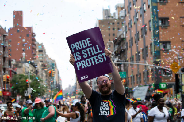An individual holds a sign that says "Pride Is Still Protest" in the middle of a crowded pride parade