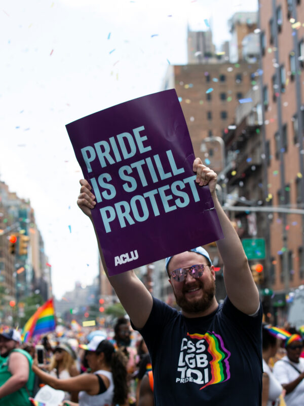 An individual holds a sign that says "Pride Is Still Protest" in the middle of a crowded pride parade