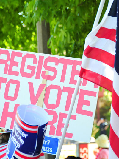 A sign reading "Register to Vote Here" next to an American flag.