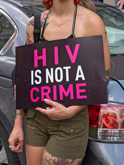 A demonstrator, leaning against a light blue car and with their face hidden holds a sign reading "HIV IS NOT A CRIME".