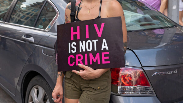 A demonstrator, leaning against a light blue car and with their face hidden holds a sign reading "HIV IS NOT A CRIME".