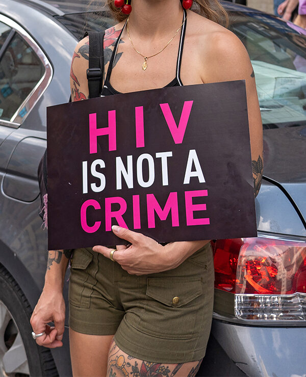 A demonstrator, leaning against a light blue car and with their face hidden holds a sign reading "HIV IS NOT A CRIME".