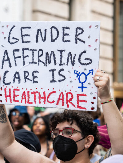 A shirtless demonstrator (wearing glasses and a black surgical mask) holds up a sign that reads" GENDER AFFIRMING CARE IS HEALTHCARE".