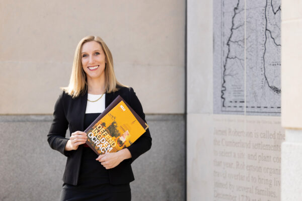 Professional woman in black suit jacket smiling while holding ACLU materials including a publication titled 'Defend Democracy' with yellow and black cover design.
