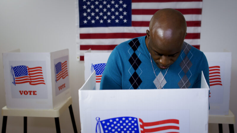 Black Man with bald head voting with American flag hung behind him.