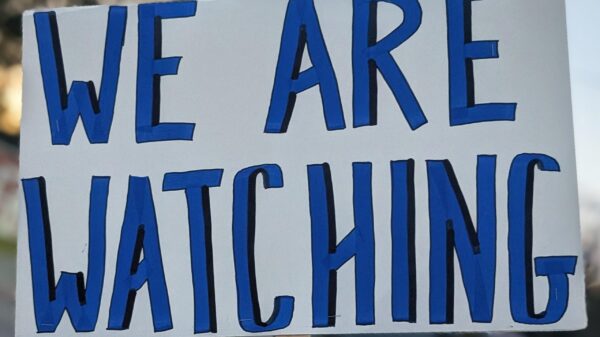 Hand holding blue and white sign that states: WE ARE WATCHING