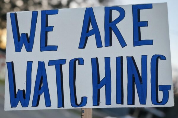 Hand holding blue and white sign that states: WE ARE WATCHING