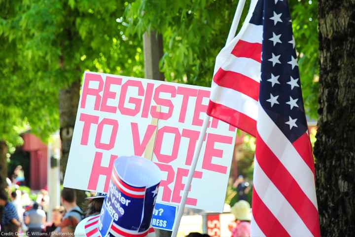 A sign reading "Register to Vote Here" next to an American flag.
