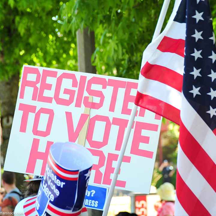 A sign reading "Register to Vote Here" next to an American flag.